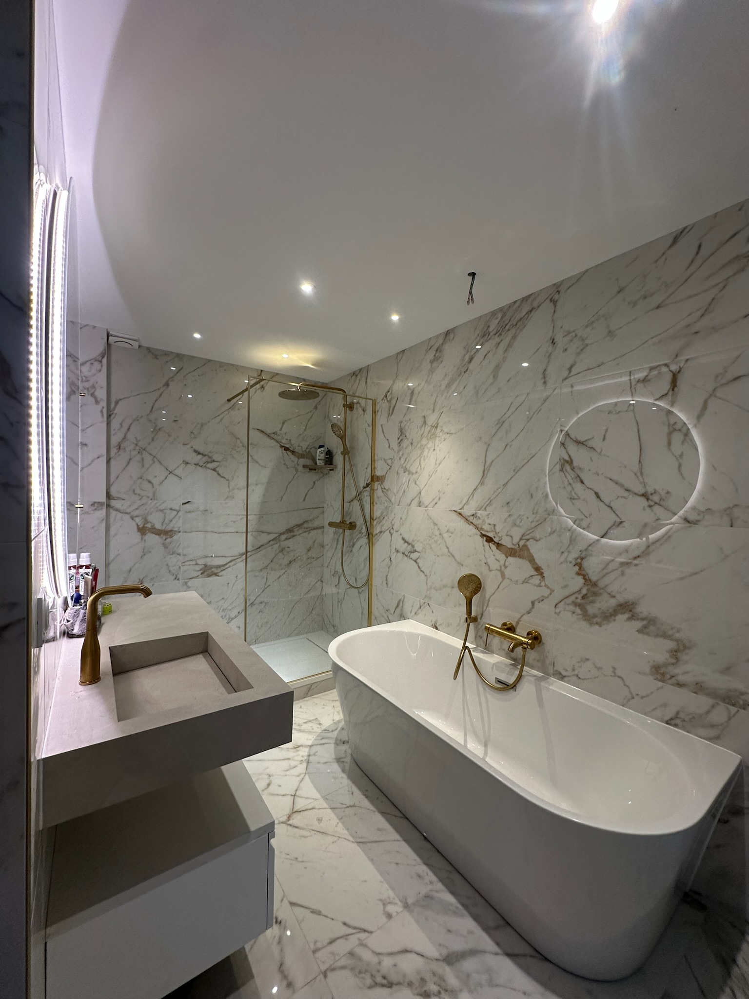 Plan de travail de cuisine avec évier rond en inox, robinet mural chromé, crédence en carreaux blancs et meubles en bois clair, éclairé par une fenêtre avec stores.