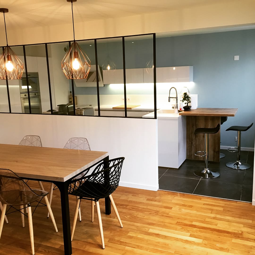 Plan de travail de cuisine avec évier rond en inox, robinet mural chromé, crédence en carreaux blancs et meubles en bois clair, éclairé par une fenêtre avec stores.